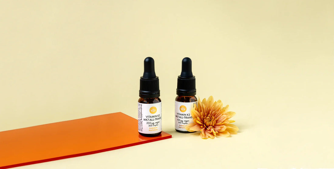 Two bottles of Vitamin K2 MK7 All-Trans drops with droppers, displayed on orange surface beside yellow flower